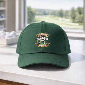 May include: A dark green baseball cap featuring a central graphic of a smiling anthropomorphic golf ball character holding a golf club. The graphic includes the text "WEEKEND WARRIOR" in a curved banner design with yellow and red accents.