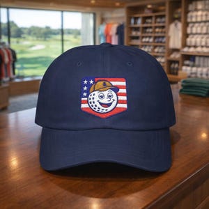 May include: A navy blue baseball cap featuring an embroidered patch of a smiling golf ball wearing a hat, set against a red, white, and blue flag design. The cap is on a wooden surface.