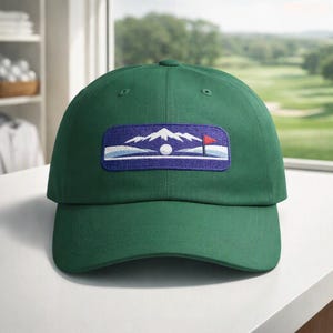 May include: A forest green baseball cap featuring a rectangular embroidered patch on the front. The patch displays a purple background with a white mountain range, a white golf ball, and a small red flag on a green putting surface.