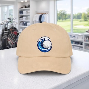 May include: A beige baseball cap with an embroidered design of a golf ball within a blue and white wave. The cap has a curved peak and a classic six-panel design, suitable for golf enthusiasts.