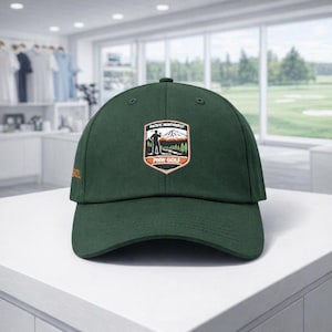 May include: A dark green baseball cap featuring a central embroidered patch with a mountain landscape, a golfer silhouette, and the text Pacific Northwest PNW Golf. The side of the cap includes orange embroidered text that reads GOLF.
