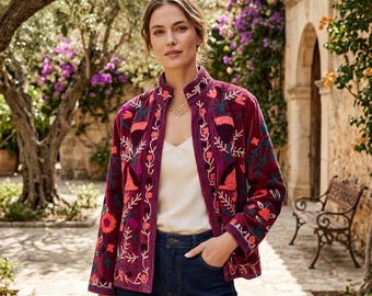 May include: A deep burgundy velvet jacket featuring a mandarin collar and long sleeves, adorned with vibrant floral embroidery in shades of orange, pink, and green. The jacket is worn over a simple cream-colored camisole and dark blue denim jeans.