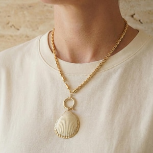 May include: A gold-toned necklace featuring a large, cream-colored seashell pendant. The pendant is detailed with ribbed lines and a gold border. The necklace has a thick chain and a circular clasp, creating a beach-inspired accessory.