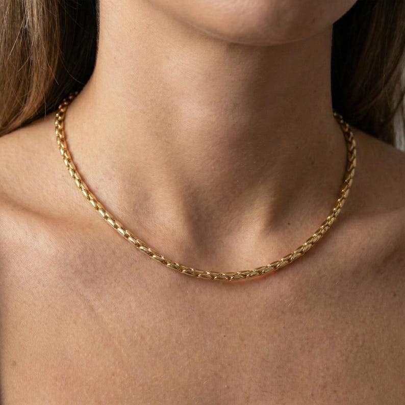May include: A gold-colored chain necklace with a textured, woven design. The necklace is worn around the neck and rests on the collarbone. The jewelry is a simple, elegant accessory.