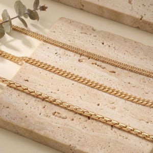 May include: Close-up of four gold-colored necklaces with different chain styles. The chains are displayed on a light beige, textured surface. A sprig of greenery is visible in the upper left corner.