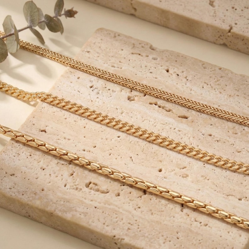 May include: Close-up of four gold-colored necklaces with different chain styles. The chains are displayed on a light beige, textured surface. A sprig of greenery is visible in the upper left corner.