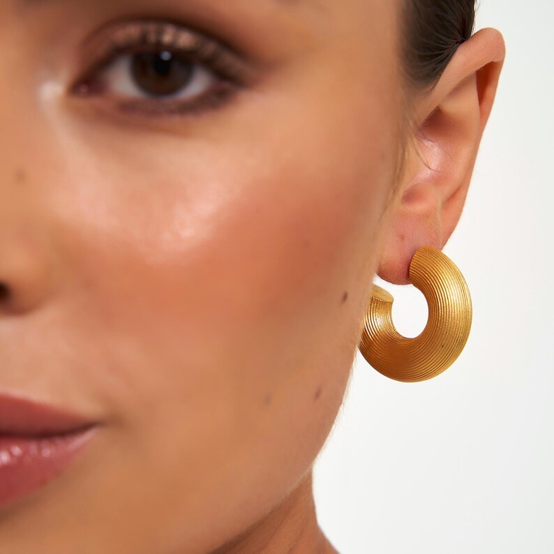 May include: Close-up of a person's face with a gold-colored, textured hoop earring. The earring is a thick, semi-circular design with a ridged surface. The skin tone is light with a subtle glow.