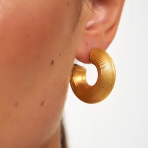 May include: Close-up of a gold-colored hoop earring. The earring has a textured, ridged surface and a rounded shape. The earring is worn on an ear, with the skin visible in the background. The earring is a bold, statement piece of jewelry.