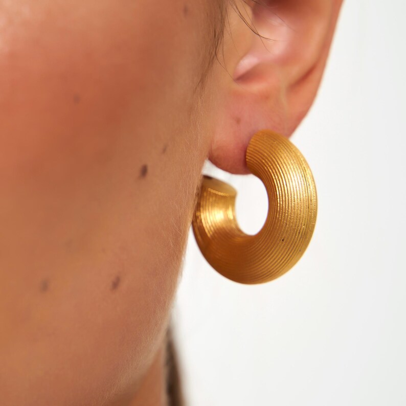 May include: Close-up of a gold-colored hoop earring. The earring has a textured, ridged surface and a rounded shape. The earring is worn on an ear, with the skin visible in the background. The earring is a bold, statement piece of jewelry.
