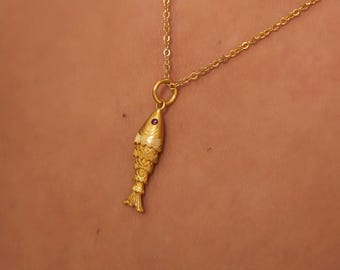 Puede incluir: Un collar con colgante de pez dorado. El colgante de pez tiene detalles intrincados y una pequeña gema redonda de color púrpura. La cadena del collar también es dorada y delicada. El collar se muestra sobre un fondo neutro.