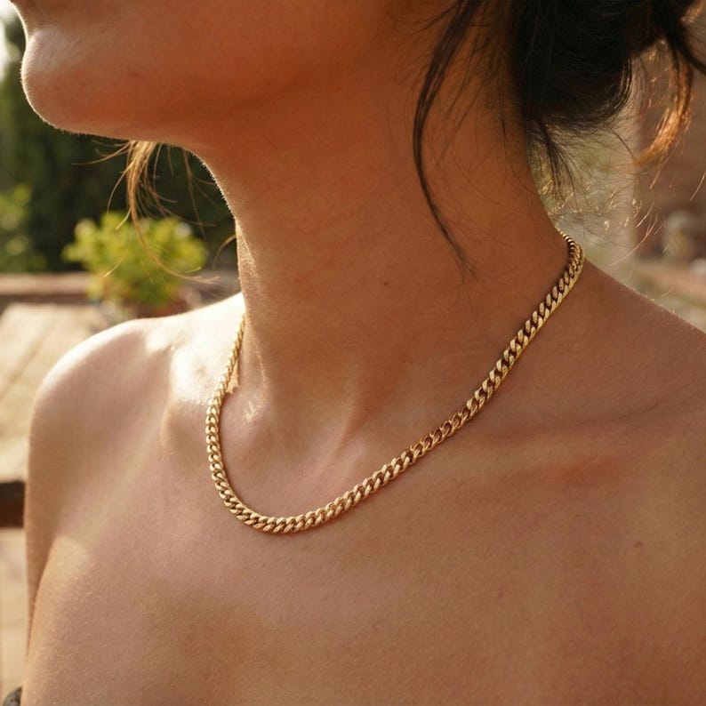 May include: A gold-colored curb chain necklace. The necklace is made of interlocking links and rests on a person's collarbone. The chain has a shiny, reflective surface, and the overall aesthetic is simple and elegant.