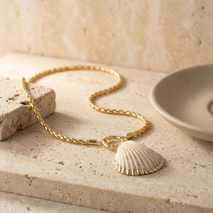 May include: Gold-toned necklace with a textured chain and a seashell pendant. The pendant is off-white with a gold-colored trim. The necklace is displayed on a light-colored, textured surface, suggesting a coastal theme.