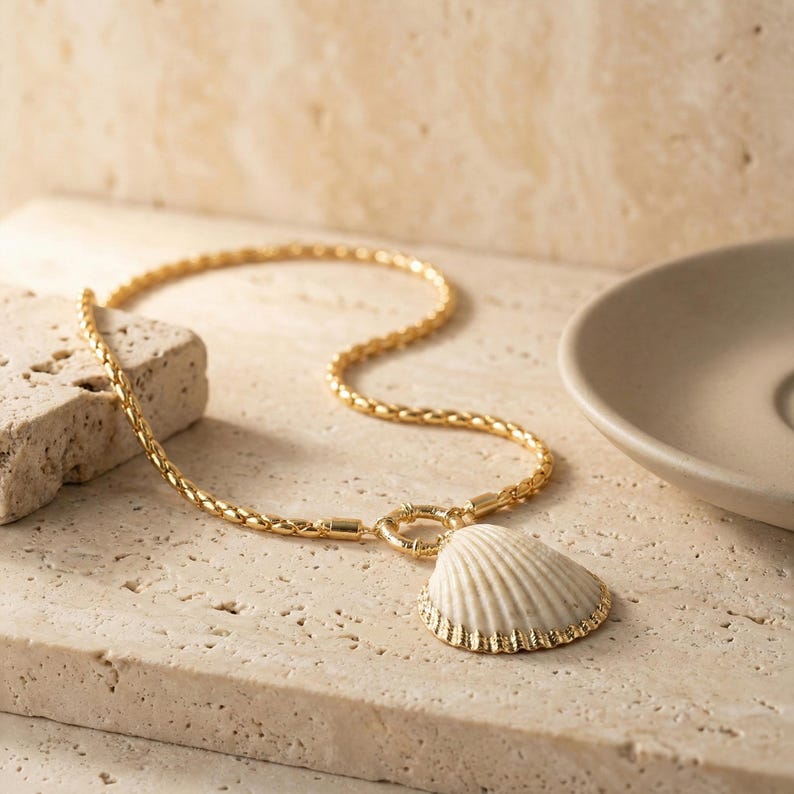 May include: Gold-toned necklace with a textured chain and a seashell pendant. The pendant is off-white with a gold-colored trim. The necklace is displayed on a light-colored, textured surface, suggesting a coastal theme.