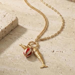 May include: Gold-toned necklace with a fish pendant, a red and white striped ball, and a small wing charm. The chain has a curb link and rope design. The necklace is displayed on a neutral-toned surface.