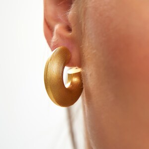 May include: A close-up of a gold-colored hoop earring. The earring has a ridged texture and a curved design. The earring is attached to an ear. The background is a soft, neutral color.