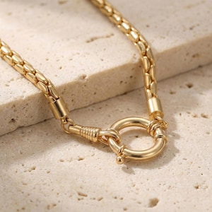 May include: A gold-colored necklace with a textured chain and a large circular clasp. The necklace is made of a shiny metal and is displayed on a light-colored surface. The clasp is a prominent feature, adding to the necklace's overall design.