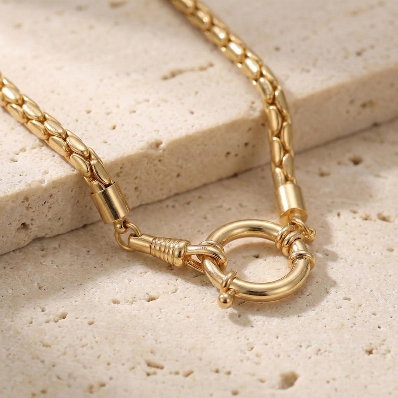 May include: A gold-colored necklace with a textured chain and a large circular clasp. The necklace is made of a shiny metal and is displayed on a light-colored surface. The clasp is a prominent feature, adding to the necklace's overall design.