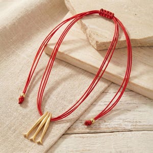 May include: A red cord necklace with a gold-colored pendant. The pendant has three stylized, elongated shapes. The necklace is displayed on a neutral-colored surface, with a light-colored background.