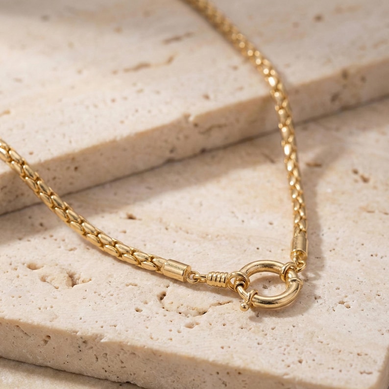 May include: A gold-colored necklace with a woven chain design. The necklace features a circular clasp and is displayed on a textured, light-colored surface. The chain is made of small, interlocking links.