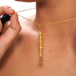 Può includere: Collana con pendente a forma di pesce dorato. Il pendente presenta un design dettagliato a forma di pesce con occhi di gemme verdi. La catena della collana è delicata e anch'essa dorata. Il pendente a forma di pesce è sospeso da un piccolo anello.