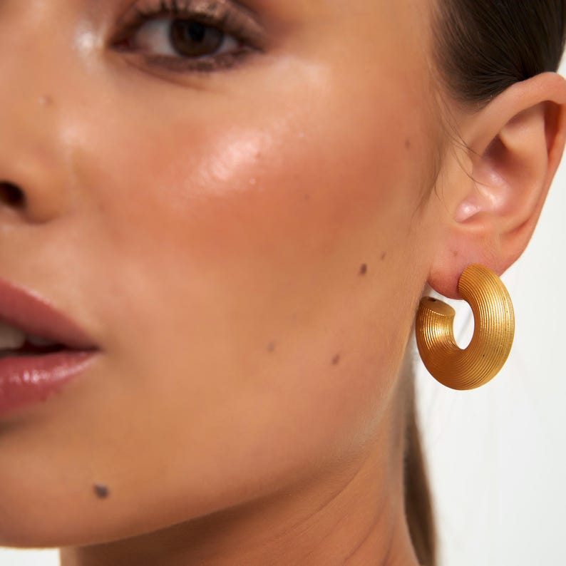 May include: Close-up of a person's face with a gold-colored, textured hoop earring. The earring is a thick, rounded shape with a ridged surface. The skin tone is light with visible freckles.