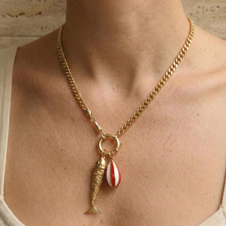 May include: Gold-toned necklace with a chunky chain. The pendant features a detailed fish charm and a red and white striped teardrop charm. The necklace is secured with a large clasp.