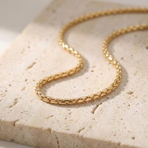May include: A gold-colored chain necklace with a textured, woven design. The necklace is displayed on a light beige, textured surface, creating a contrast that highlights the jewelry's details. The chain is made of small, interconnected links.