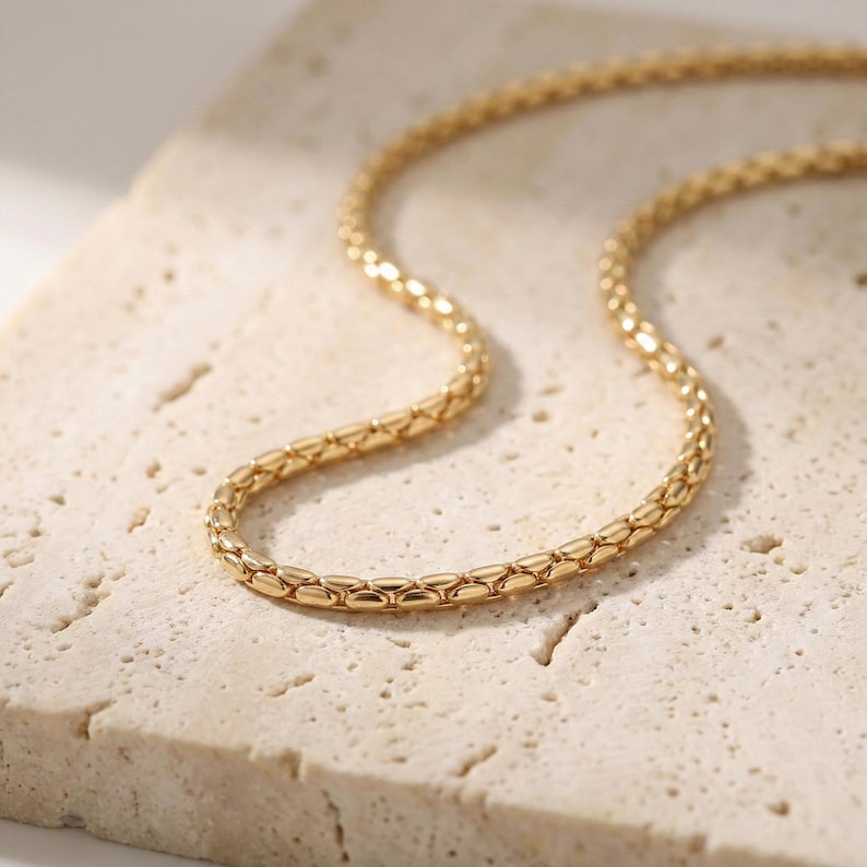 May include: A gold-colored chain necklace with a textured, woven design. The necklace is displayed on a light beige, textured surface, creating a contrast that highlights the jewelry's details. The chain is made of small, interconnected links.