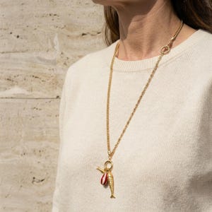 May include: A gold-colored necklace with a fish pendant. The pendant has a red and white striped element and a small gold-colored fish. The necklace is worn over a cream-colored sweater.