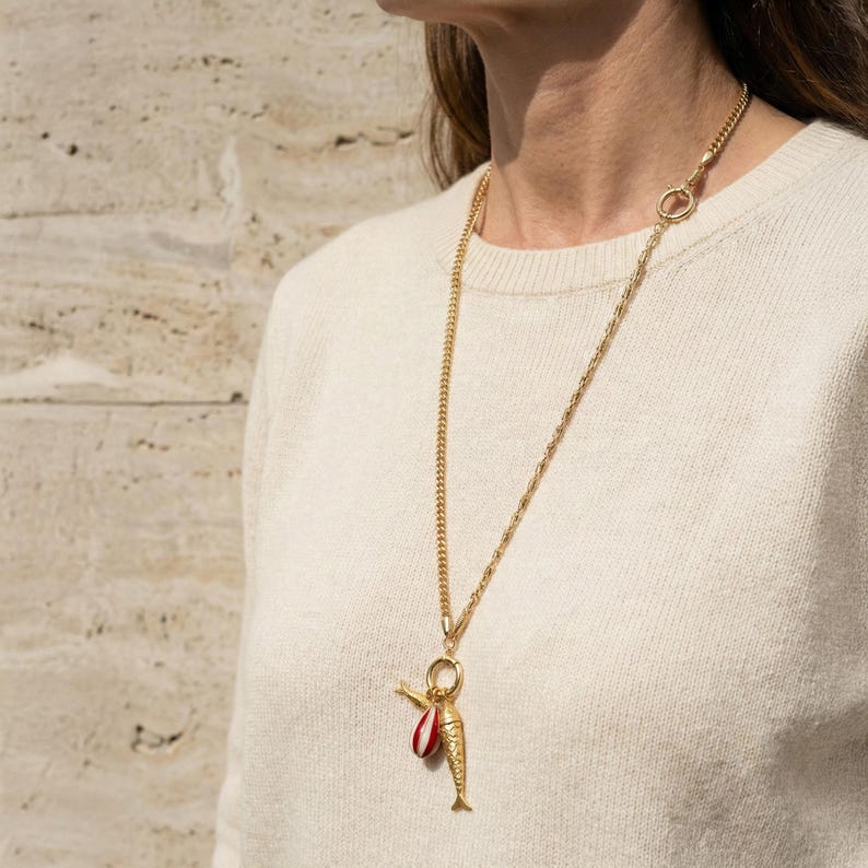 May include: A gold-colored necklace with a fish pendant. The pendant has a red and white striped element and a small gold-colored fish. The necklace is worn over a cream-colored sweater.