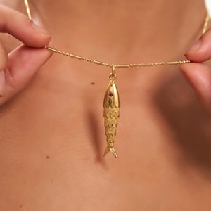 Op de afbeelding: Een goudkleurige vishangerketting met een kleine rode edelsteen oog. De vis heeft gedetailleerde schubbenpatronen en een kleine staart. De ketting heeft een delicate gouden kleur.
