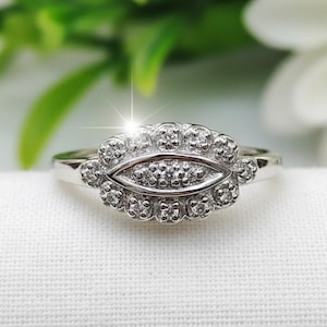 May include: A silver ring with a central oval design, surrounded by small, sparkling stones. The ring's band is thin and polished, and the stones catch the light, creating a brilliant display. The ring is set against a white background.