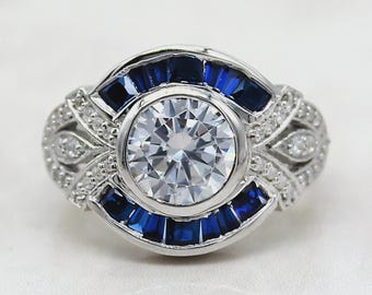 May include: A silver ring with a large, round, clear center stone. The ring features a band of small, blue rectangular stones and smaller clear stones. The design is symmetrical and detailed, with a vintage or art deco style.