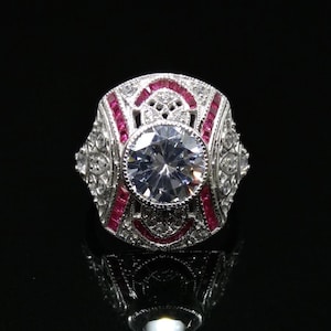 May include: A silver-toned ring with a large, clear, round center stone. The ring features red accent stones and intricate filigree detailing. The design is symmetrical and set against a black background, highlighting the ring's sparkle.