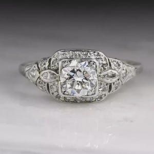 May include: A vintage-style engagement ring with a large, round-cut diamond center stone. The diamond is set in a square halo of smaller diamonds, with additional diamond accents on the band. The ring is made of white metal.