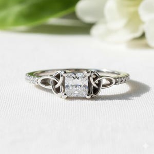 May include: A silver ring with a square, clear gemstone at the centre. The ring features a Celtic knot design on either side of the gemstone and small, clear stones along the band. The ring is set against a white background.