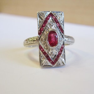 May include: A rectangular silver-toned ring featuring a central oval-cut red ruby surrounded by a geometric frame of smaller red rubies and clear stones. The metal band displays intricate engraved detailing along the sides of the ring setting.