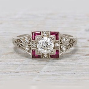May include: A vintage-style ring with a large, round, clear center stone, surrounded by smaller clear stones and square, deep red stones. The band is silver-toned with intricate filigree detailing. The ring is set against a light wood grain background.
