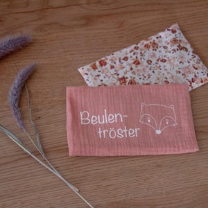 Könnte beinhalten: Ein pfirsichfarbener, rechteckiger Stoff mit dem Text "Beulen-tröster" und einer Fuchsgrafik. Ein Stoff mit Blumenmuster und getrocknetes, lavendelfarbenes Gras sind ebenfalls auf einer Holzoberfläche vorhanden.
