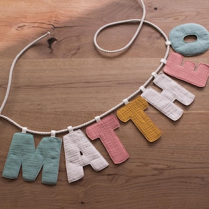 Puede incluir: Un cartel hecho a mano con la palabra "MATTHEW" en colores pastel. Las letras son de tela texturizada y est&aacute;n ensartadas en una cuerda blanca. El cartel est&aacute; expuesto sobre una superficie de madera.