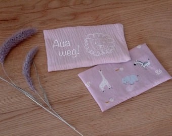 Puede incluir: Dos paños rectangulares rosa claro para bebés con ilustraciones de animales y el texto "Aua weg!". Uno presenta un león, el otro una jirafa, una cebra y un hipopótamo. Flores moradas secas sobre una superficie de madera.