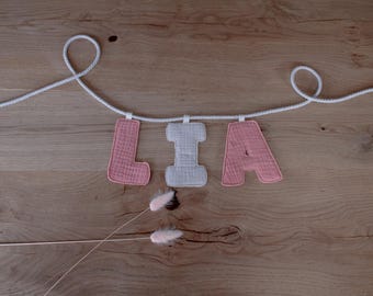 Puede incluir: Una guirnalda decorativa con las letras "LIA" en tela rosa y blanca, colgada de una cuerda blanca. Las letras están sobre un fondo de madera, con dos ramitas de flores secas debajo.