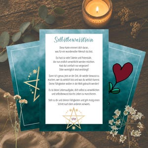 May include: Three teal and white cards with gold accents and text. The center card has a red heart illustration. The text on the cards is in German. A lit candle and dried flowers are in the background. The cards are on a wooden surface.