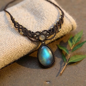 Könnte beinhalten: Eine handgefertigte Makramee-Halskette mit einem tropfenförmigen Labradorit-Anhänger mit blau-grünen Irideszenz. Die Halskette hat eine dunkelbraune Kordel mit goldfarbenen Perlen und einen runden, hellfarbigen Steinakzent.