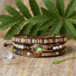Könnte beinhalten: Drei Wickelarmbänder mit braunen Lederriemen. Das obere Armband hat rechteckige Perlen in Grün, Blau und Gold. Das mittlere Armband hat einen tropfenförmigen Anhänger. Das untere Armband hat kleine, runde, farbige Perlen.