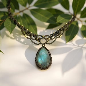 Puede incluir: Collar de macramé hecho a mano con un colgante de labradorita en forma de lágrima con tonos iridiscentes azules y verdes. El collar tiene un cordón marrón oscuro con detalles dorados y una pequeña piedra blanca redonda.