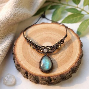 Peut inclure: Un collier macramé artisanal avec un pendentif en pierre précieuse bleu-vert irisé en forme de goutte. Le collier est composé d'un cordon tissé marron foncé avec des perles dorées et une petite pierre ronde. Le collier est présenté sur une tranche de bois.