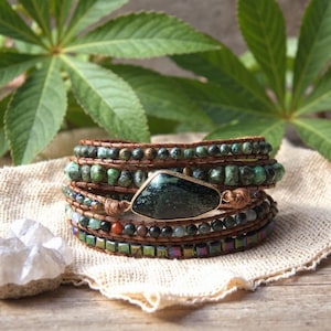 Puede incluir: Pulsera envolvente de varias hebras con cuentas verdes y marrones, con una gema grande de color verde oscuro en un engaste dorado. La pulsera está hecha con cuero y piedras naturales, ideal para un estilo bohemio, y mide unos 18 cm de contorno.