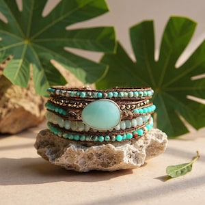 May include: A multi-strand wrap bracelet with turquoise and gold beads, and a large oval turquoise stone. The bracelet is wrapped with brown leather cord. The bracelet is displayed on a light-coloured stone, with green leaves in the background.