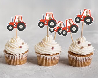 May include: Three cupcakes with white frosting and decorative sprinkles. Each cupcake is topped with a red tractor-shaped pick featuring a baby and the number 2. The cupcakes are on a light gray surface.